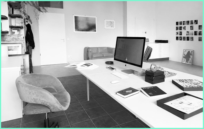 Arbeitsplatz im Atelier: Schreibtisch mit Rechner, aufgeschlagenen Büchern und gerahmten Arbeiten, dahinter der offene Raum.
