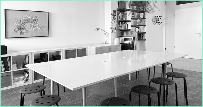 Blick ins Atelier: der lange helle Arbeitstisch mit Hockern, dahinter Regale, Zeichenschrank und eine gerahmte Arbeit.